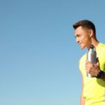 Young sporty man with water bottle against blue sky on sunny day. Space for text