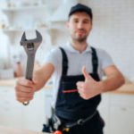 Young plumber hold wrench in hand. He show it to camera and hold big thumb up. Guy stand in kitchen. White background. Daylight.