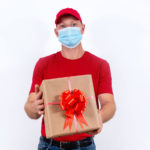 Safe delivery of gifts for holidays. A courier in red uniform and protective medical mask holds box with a bow. Contactless remote gift orders in quarantine during the coronavirus pandemic.