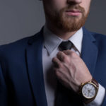 portrait of a young bearded handsome guy in a business suit, looking at the camera, demonstrating a wristwatch, with a leather strap. Advertising men’s watches. On a gray background.