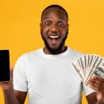 Excited African Guy Showing Smartphone Holding Money Over Yellow Background